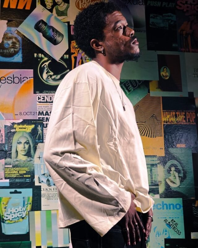 A man in a loose cream shirt standing in front of a wall papered edge to edge with magazine covers and gig posters.