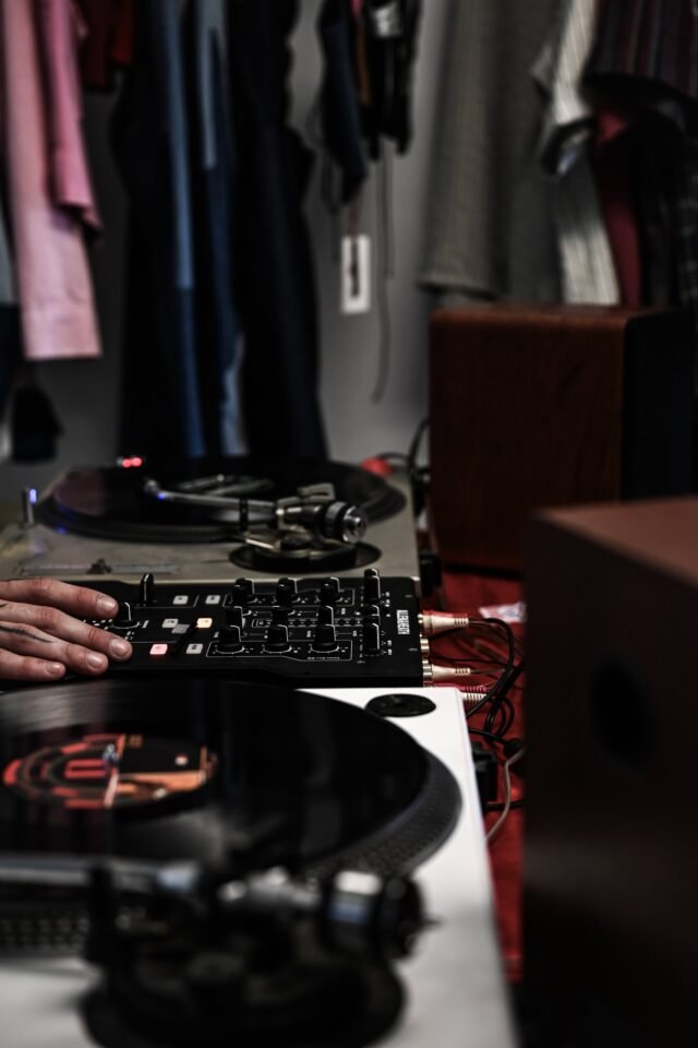 Turntables and a mixer in low light, one hand resting on the controls, a rail of clothes behind.