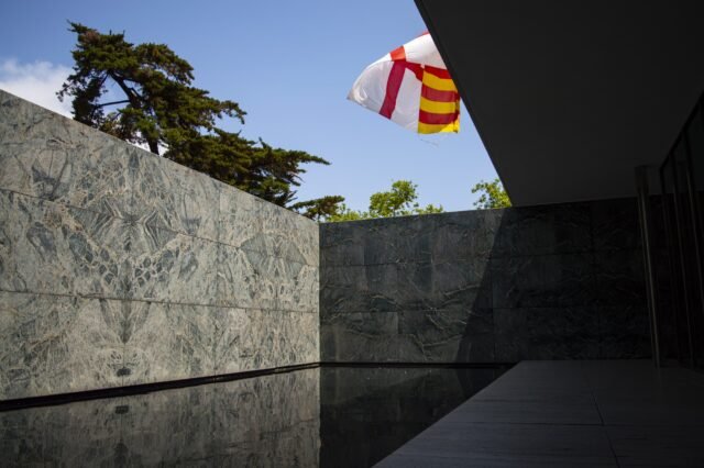 A flag lifting in the wind above the green marble walls and still black reflecting pool of a modernist pavilion.