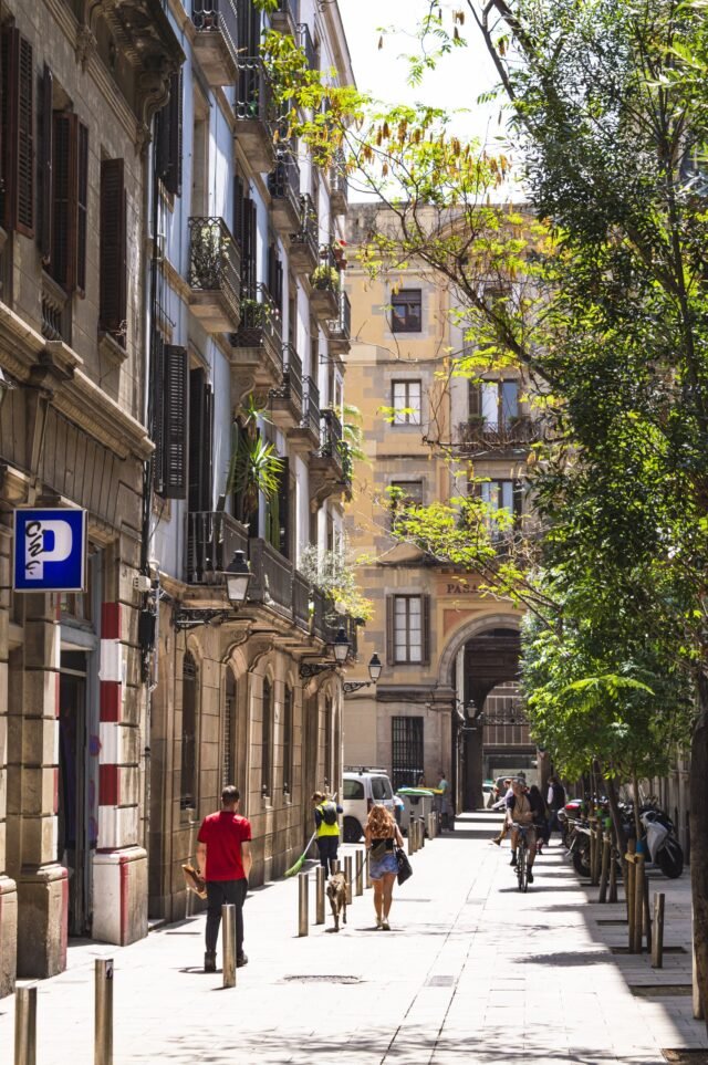 A narrow street between old apartment blocks, trees along one side and people walking into the distance.