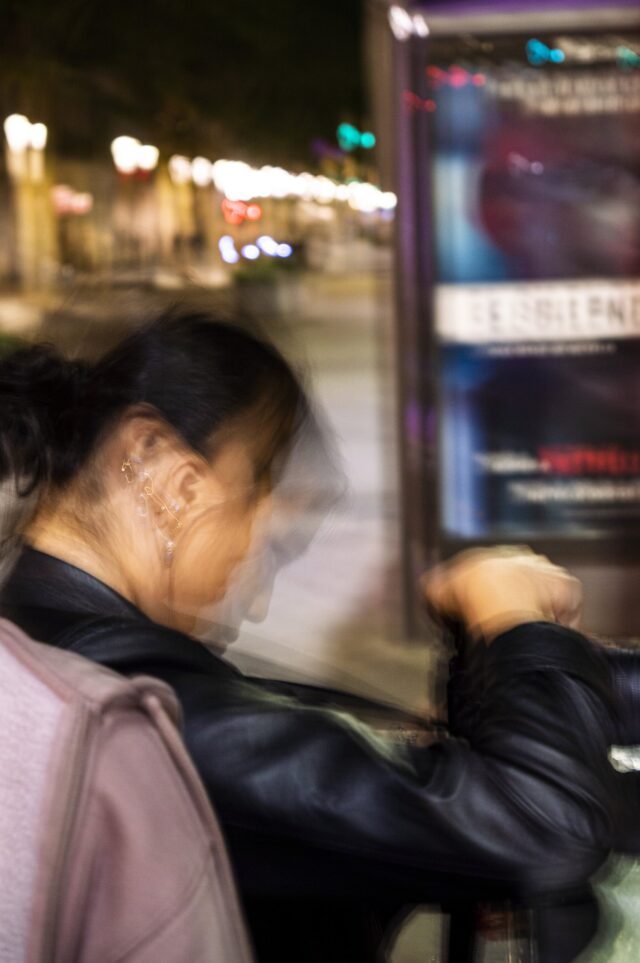A woman leaning against a lit advertising panel on a night street, caught mid-movement.