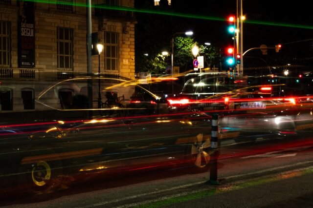 A city crossroads at night, traffic pulled into red and gold streaks past a lit stone facade.