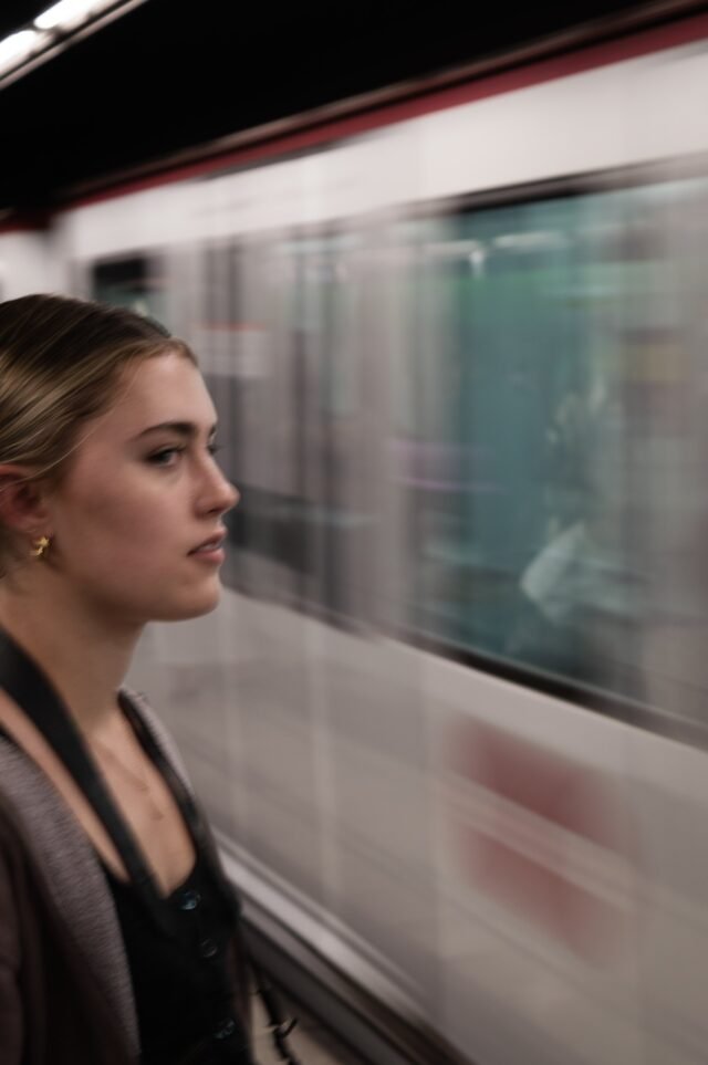 A young woman in profile on a platform while a train rushes past behind her, pulled into a blur.