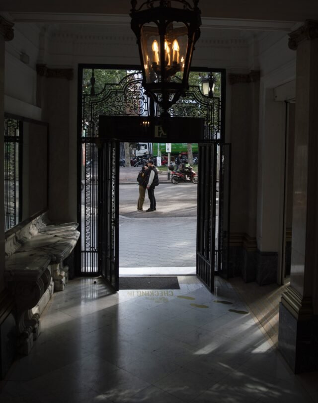 Two people embracing on the pavement, seen from inside a marble entrance hall through open wrought-iron doors.