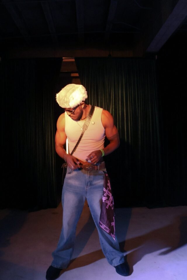 A man in a white vest, cap and printed jeans, lit against a dark curtain.