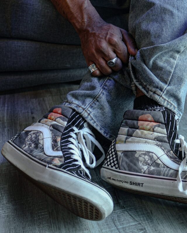 Close on a pair of printed high-top trainers and turned-up jeans, a ringed hand resting on the knee.