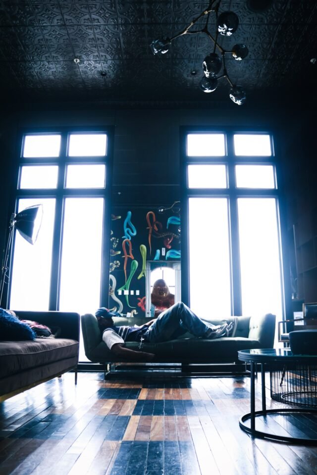 A person lying along a low sofa between two tall windows, in a room with a dark ceiling and a wooden floor.